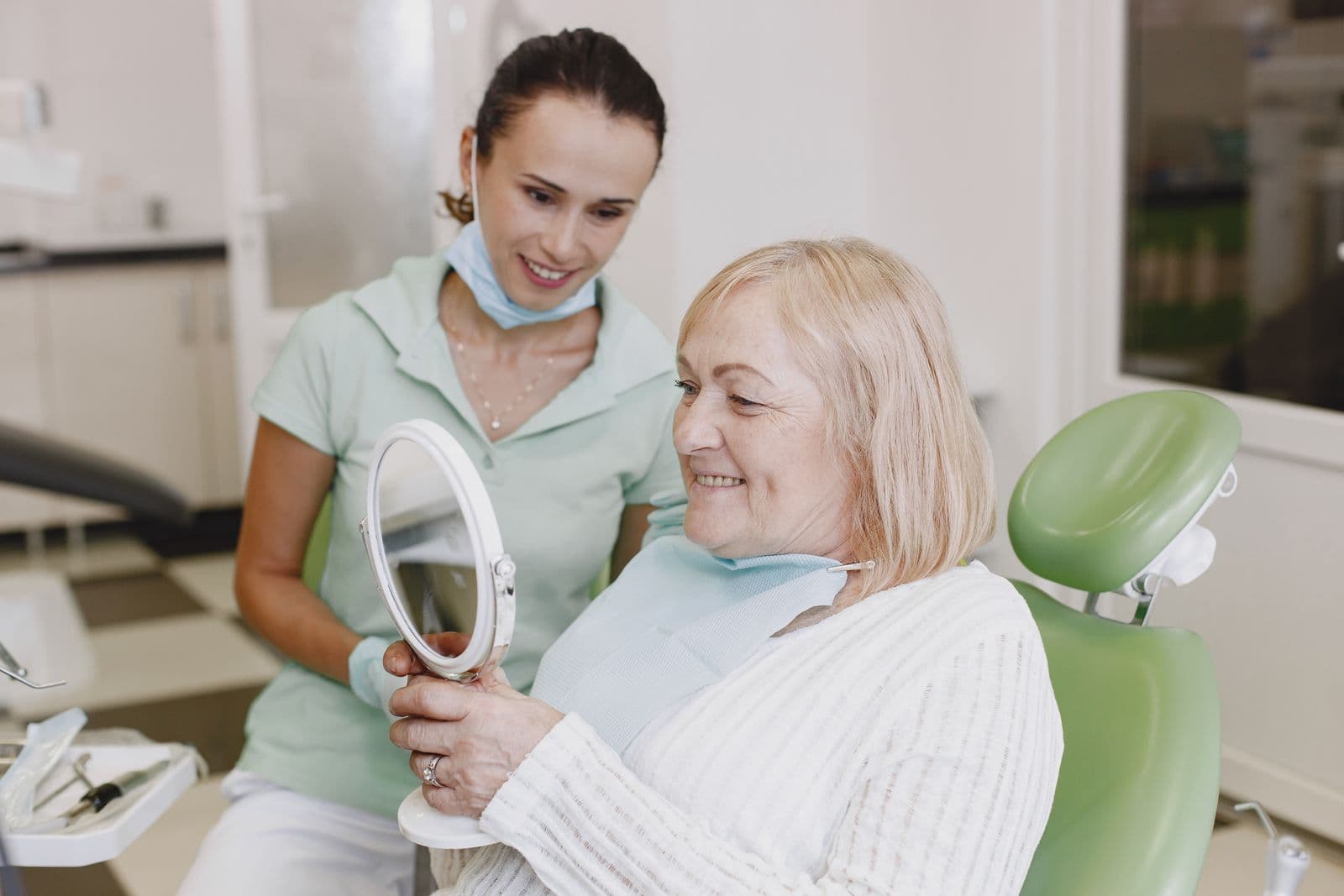 Patient seated in a dental chair holding a hand mirror, a dentist beside her